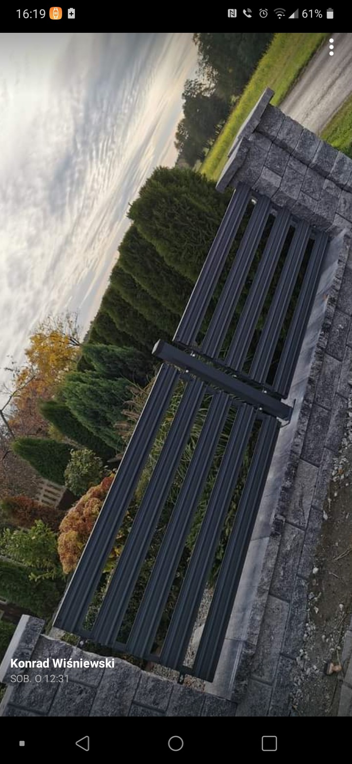 Nowoczesna, metalowa brama posesji w Krzepicach, z pionowymi profilami, osadzona na kamiennych słupkach i podmurówce, na tle zieleni i pochmurnego nieba.