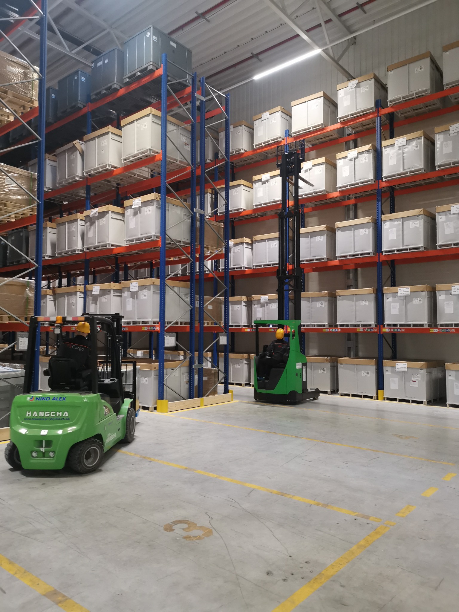 Wnętrze magazynu z regałami wypełnionymi białymi i szarymi kontenerami, dwa zielone wózki widłowe HANGCHA na betonowej podłodze z żółtymi liniami.