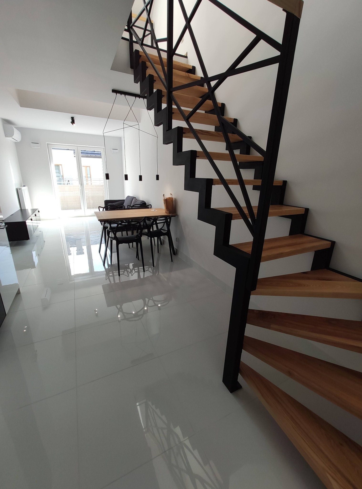 Nowoczesne wnętrze: białe ściany, lśniąca podłoga, drewniane schody z czarną metalową konstrukcją, stół z krzesłami i minimalistyczna lampa wisząca.