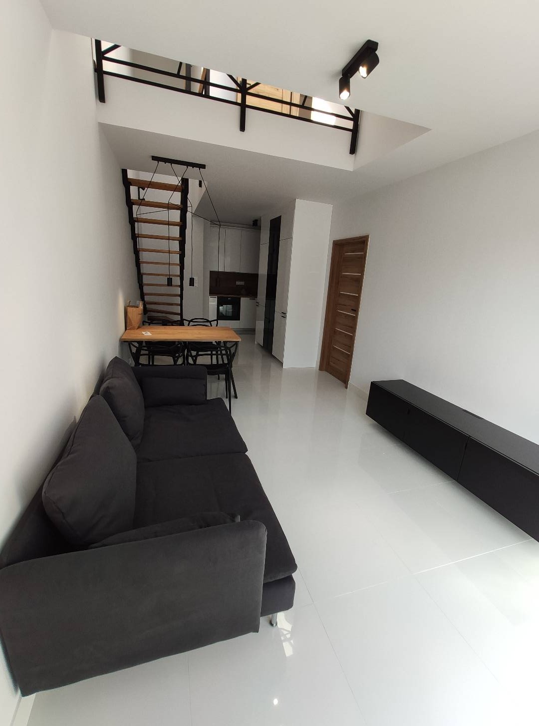 Nowoczesne wnętrze po remoncie: białe ściany, podłoga z połyskiem, antresola ze schodami, szara sofa i czarne meble. Minimalistyczny design.