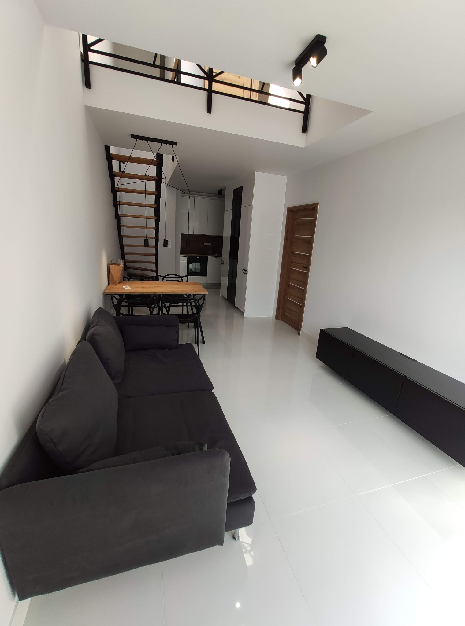 Nowoczesne wnętrze: szara sofa, białe płytki, drewniane schody i stół. Minimalistyczny design z czarnymi akcentami oświetlenia i mebli. Jasne, przestronne pomieszczenie.