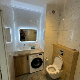 OLEKSII BIESIEDA - Nowoczesna łazienka z umywalką nablatową, lustrem z podświetleniem LED, toaletą podwieszaną w obudowie z drewnopodobnych paneli i pralką pod blatem, wykończona płytkami imitującymi marmur.