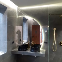 OLEKSII BIESIEDA - Nowoczesna łazienka z szarymi płytkami imitującymi marmur, podświetlanym lustrem, dwiema czarnymi umywalkami nablatowymi z złotymi bateriami i złotym zestawem prysznicowym; w lustrze odbija się...