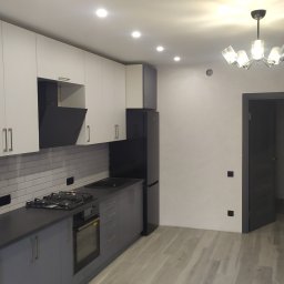 OLEKSII BIESIEDA - Nowoczesna kuchnia z białymi szafkami górnymi i szarymi dolnymi, czarnym okapem i lodówką, jasnymi płytkami imitującymi cegłę nad blatem, szarą podłogą imitującą drewno oraz dekoracyjną lampą...