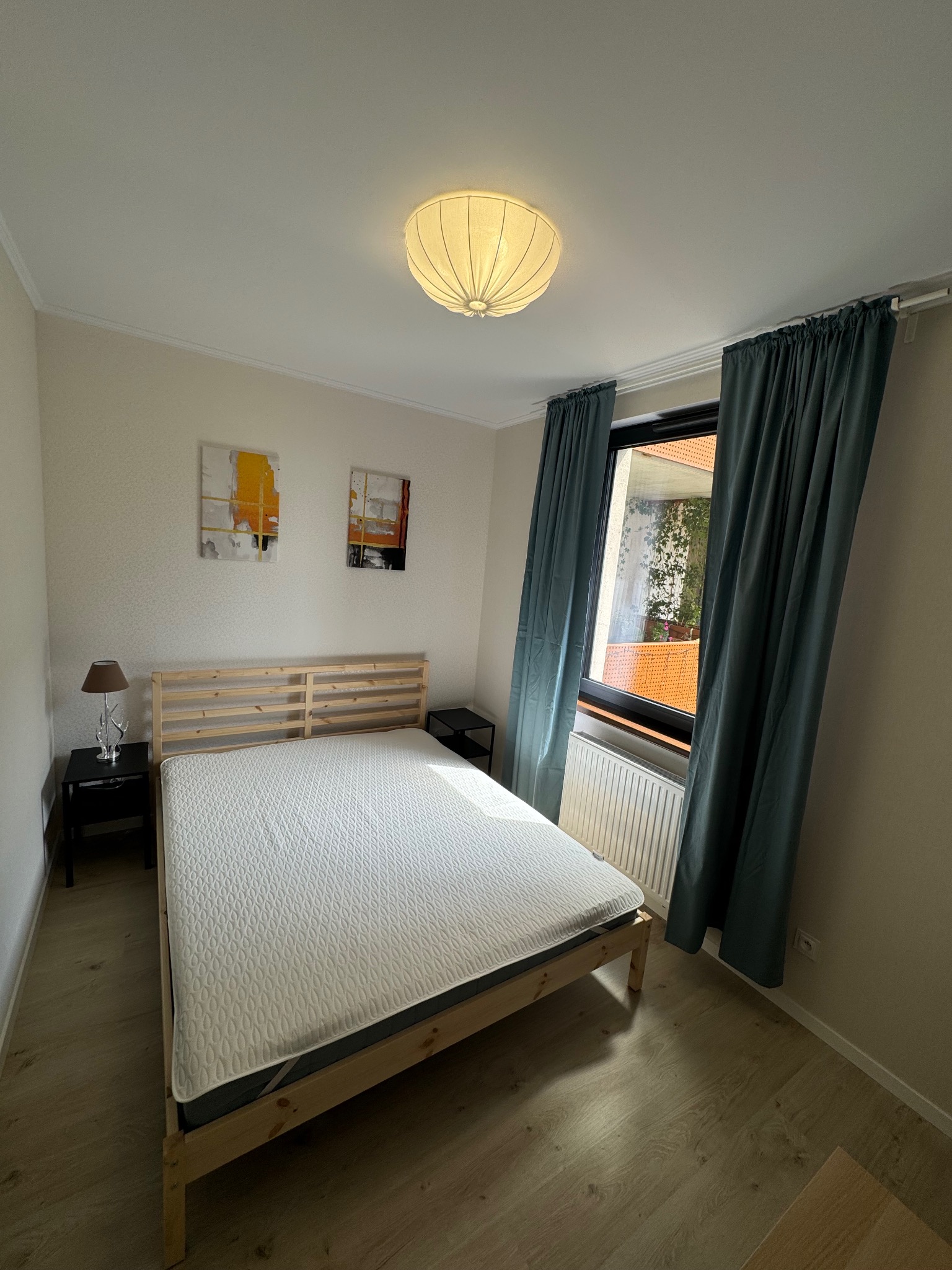 Jasna sypialnia z drewnianym łóżkiem, wzorzystą pościelą, lampką nocną i abstrakcyjnymi obrazami na ścianie, okno zasłonięte turkusowymi zasłonami.