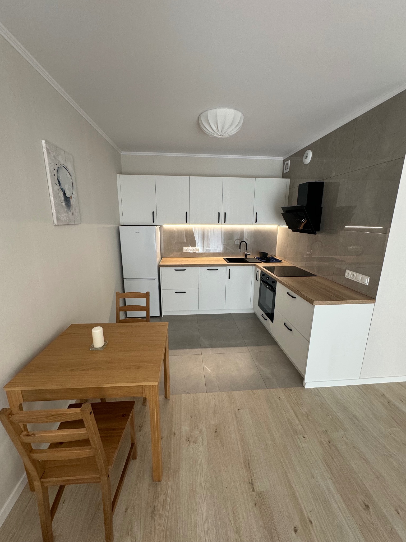 Jasna kuchnia z białymi szafkami, drewnianym blatem i czarnym okapem, widok na drewniany stół z krzesłami i świecą.