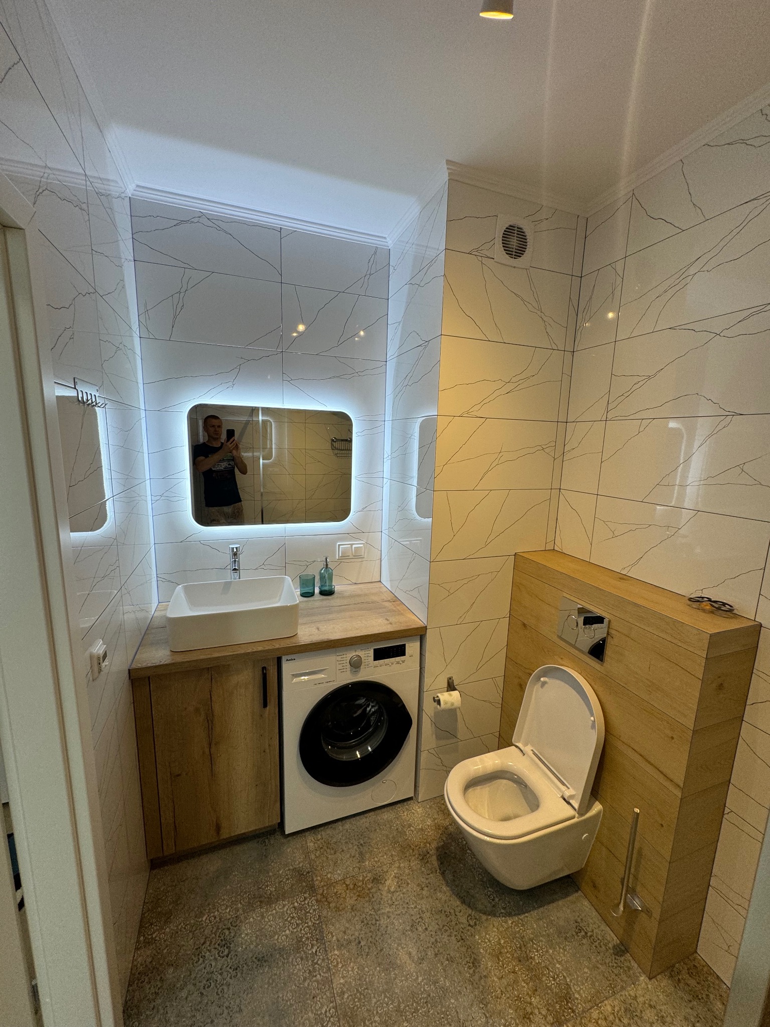 Nowoczesna łazienka z umywalką nablatową, lustrem z podświetleniem LED, toaletą podwieszaną w obudowie z drewnopodobnych paneli i pralką pod blatem, wykończona płytkami imitującymi marmur.