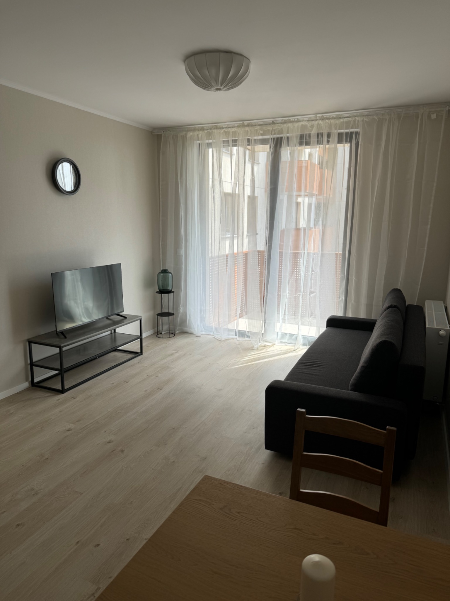 Jasne wnętrze pokoju dziennego z drewnianą podłogą, szarą sofą, telewizorem na czarnym stoliku, okrągłym lustrem na ścianie i zasłonami w oknie balkonowym.
