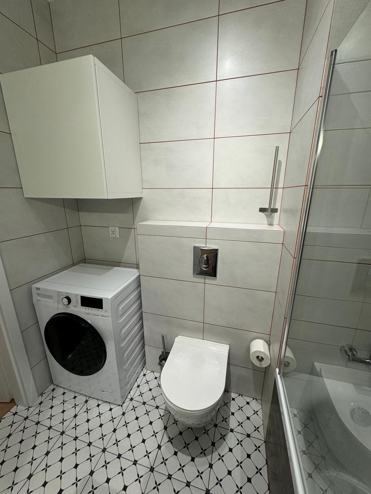 Nowoczesna łazienka z białą pralką obok podwieszanej toalety, białe płytki z czerwoną fugą i wzorzysta podłoga.