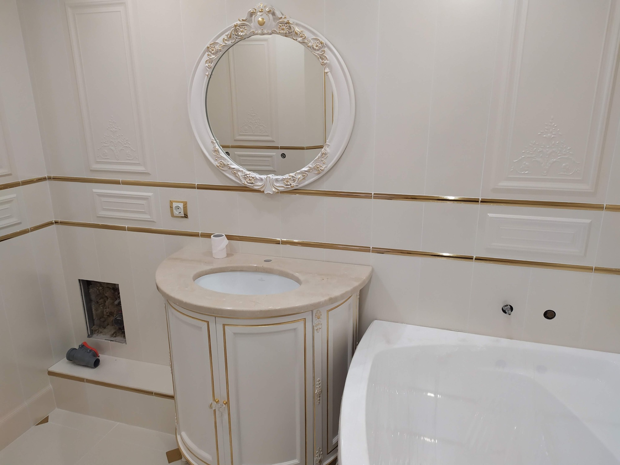 Elegancka łazienka w stylu klasycznym z białymi płytkami i złotymi akcentami, owalne lustro z ozdobną ramą, szafka pod umywalką z marmurowym blatem, widoczna instalacja wodna w ścianie