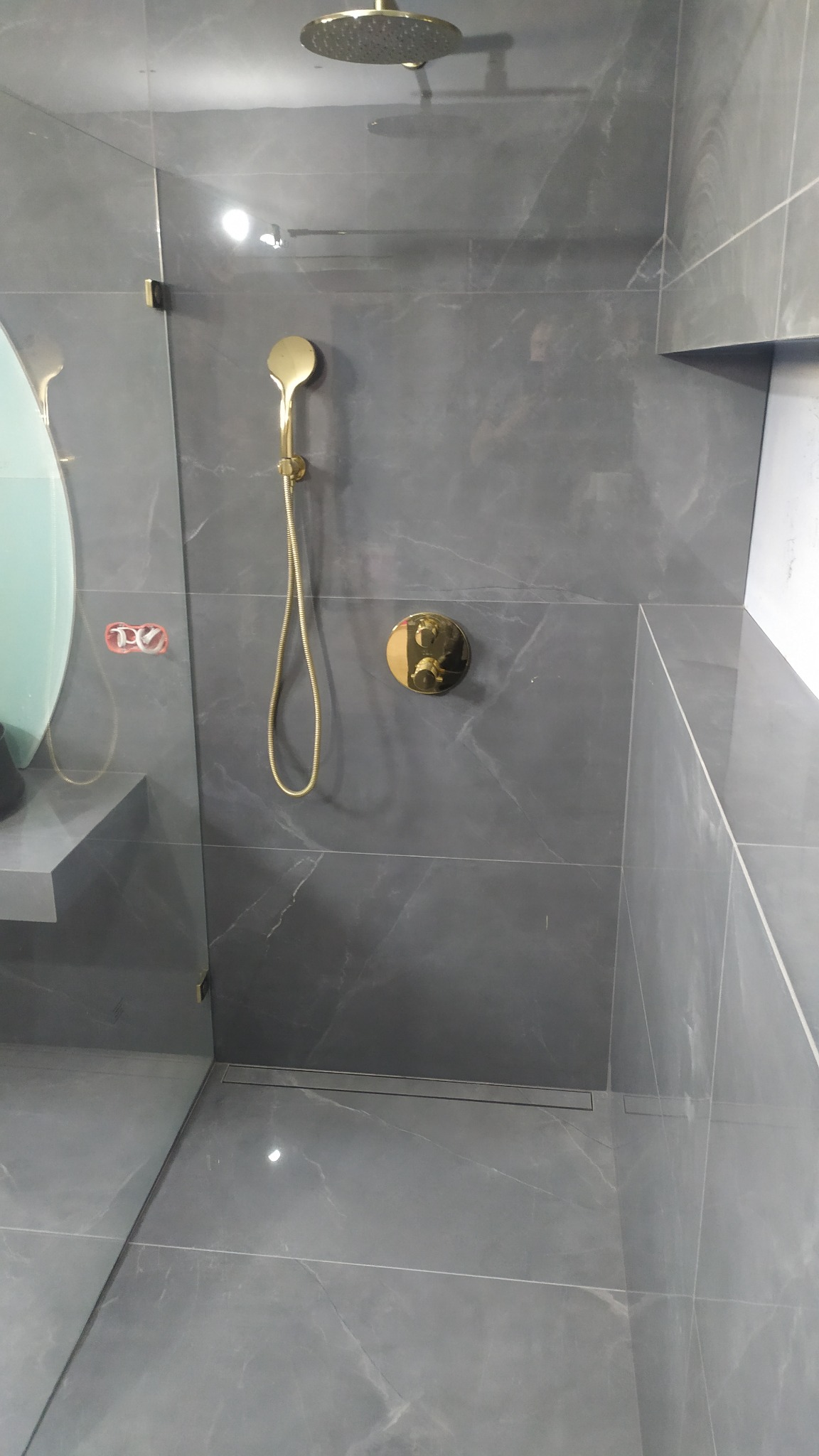 Szara kabina prysznicowa z marmurowymi płytkami, złotą deszczownicą i baterią, widoczny odpływ liniowy w podłodze.