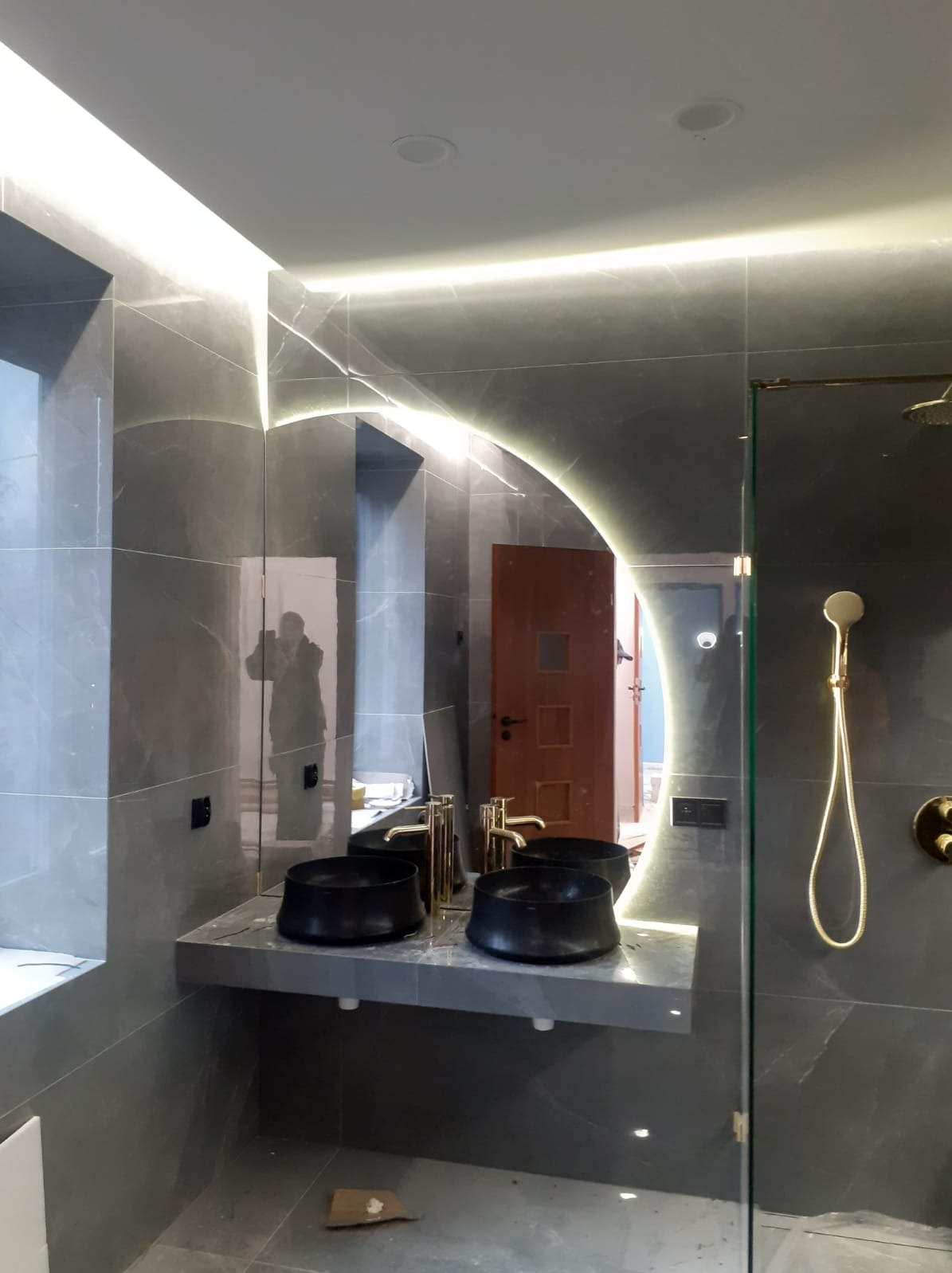 Nowoczesna łazienka z szarymi płytkami imitującymi marmur, podświetlanym lustrem, dwiema czarnymi umywalkami nablatowymi z złotymi bateriami i złotym zestawem prysznicowym; w lustrze odbija się...