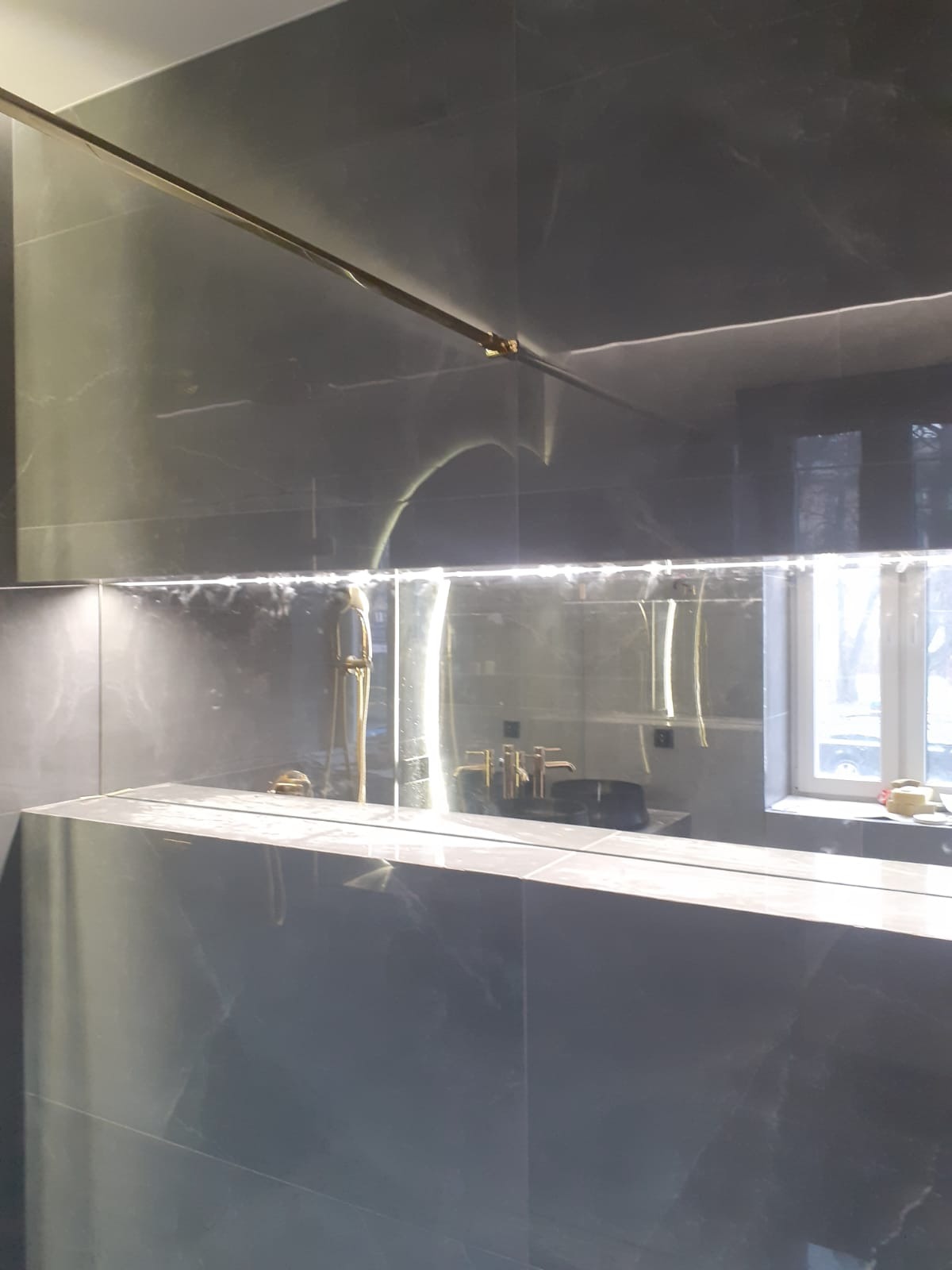 Szare marmurowe płytki w łazience z podświetlanym lustrem i złotą armaturą, widoczne odbicie okna.
