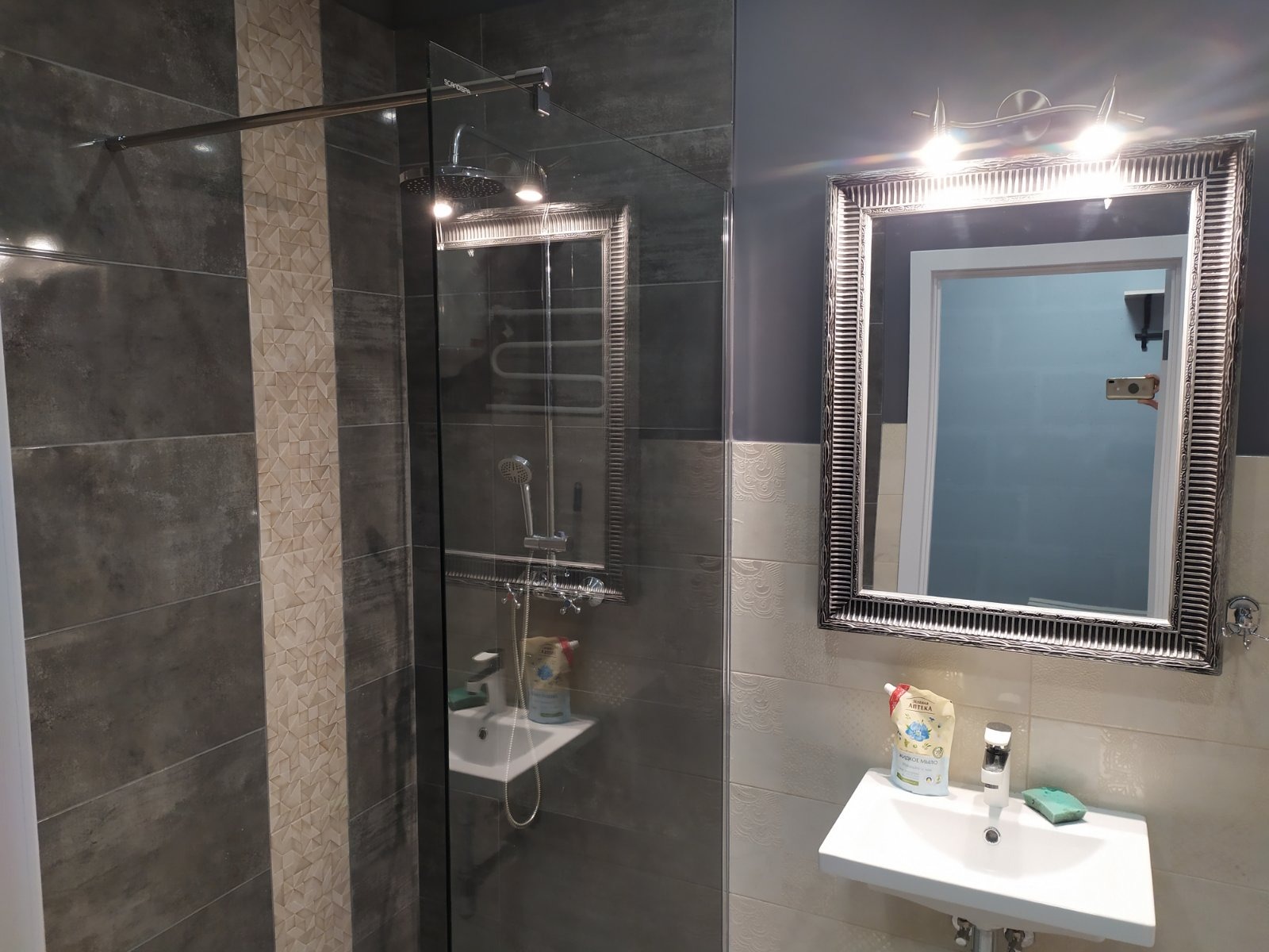 Nowoczesna łazienka z szarą glazurą, kabiną prysznicową typu walk-in z deszczownicą i lustrem w ozdobnej ramie, oświetlonym dwoma punktowymi lampami.