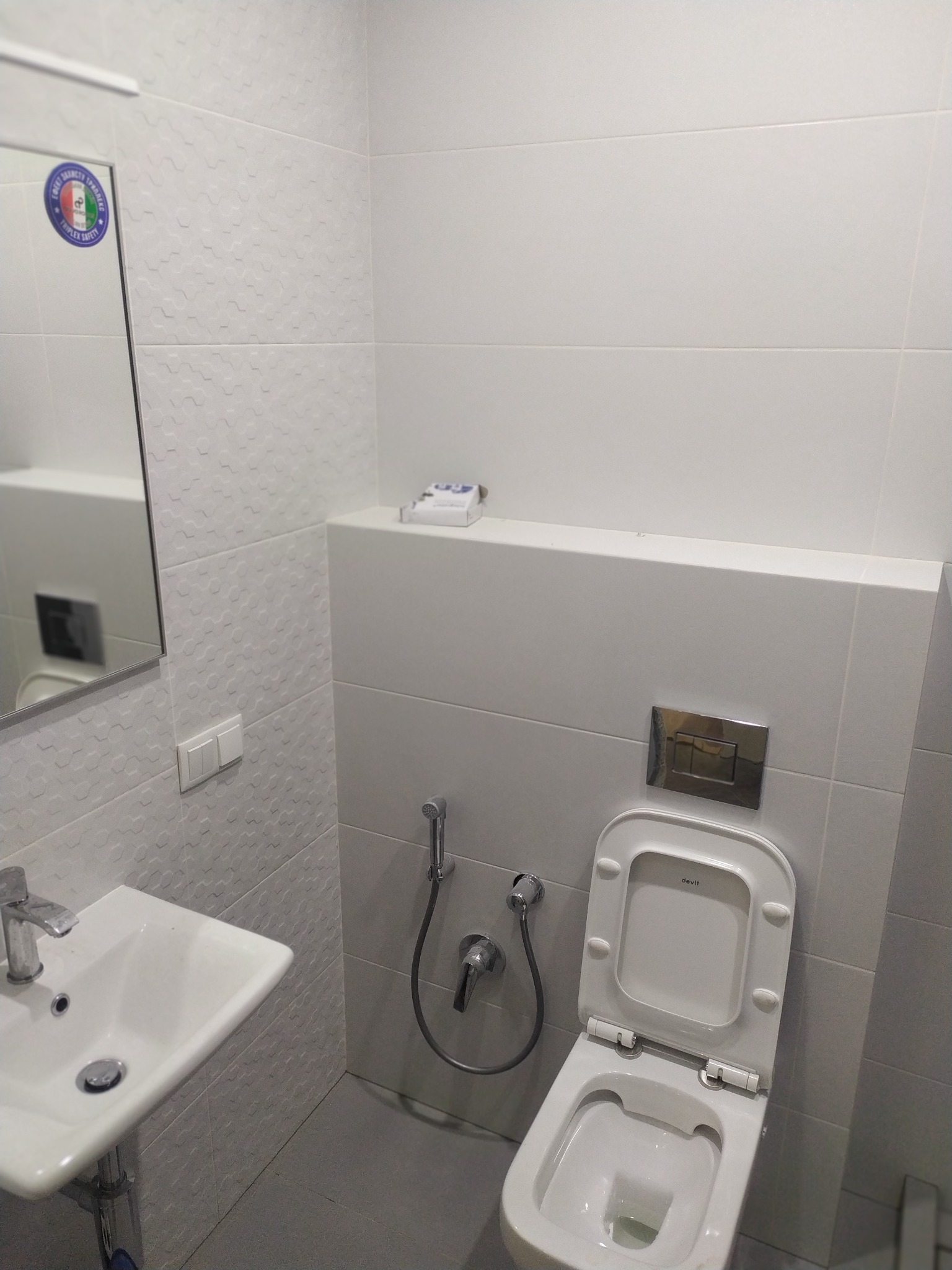 Biała łazienka po renowacji z umywalką, toaletą z bidetką i lustrem, wykończona płytkami o subtelnym wzorze.