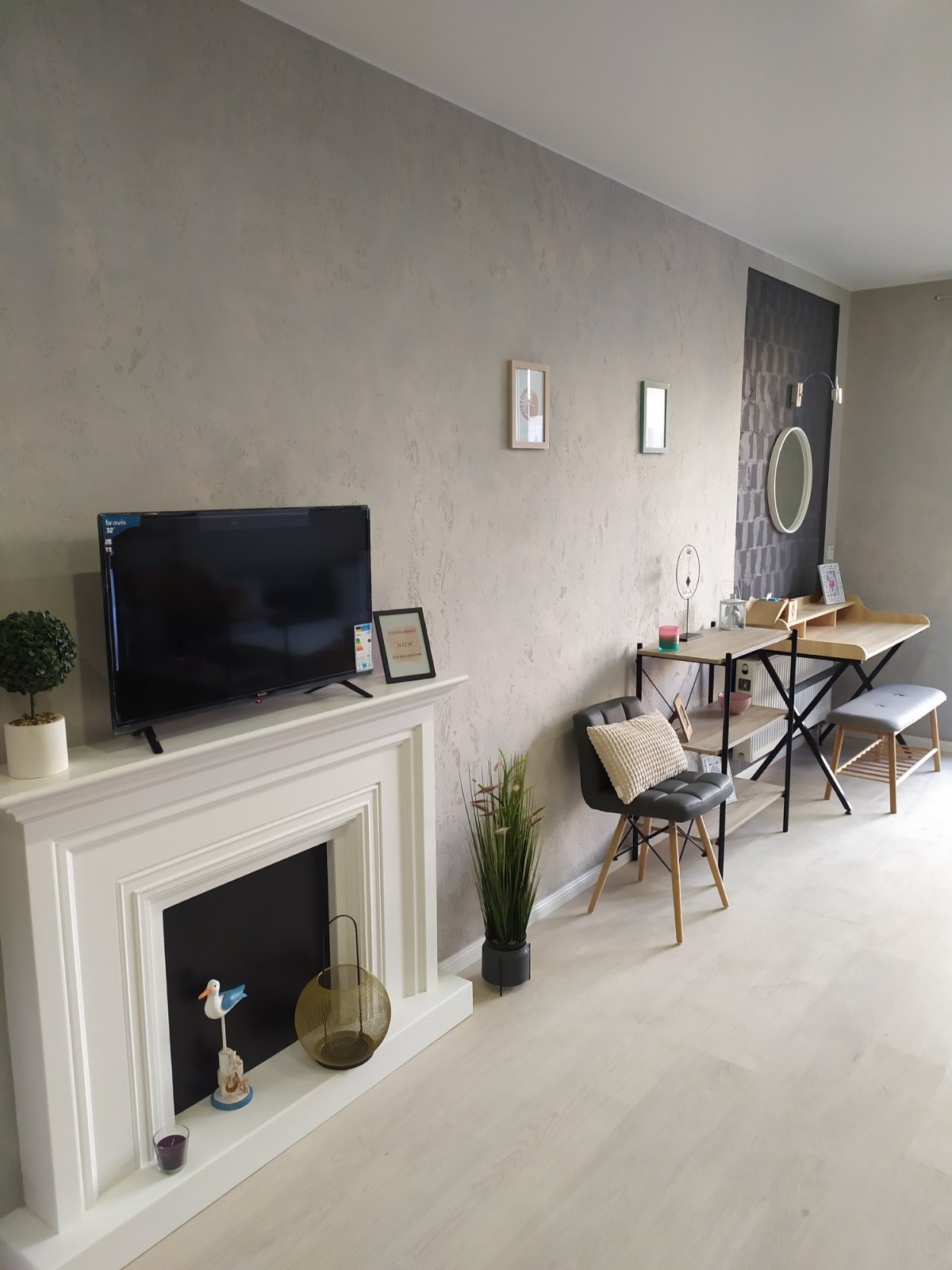 Jasne wnętrze pokoju z dekoracyjną ścianą o fakturze betonu, białym kominkiem z ozdobami, biurkiem i krzesłem, oraz jasną podłogą.