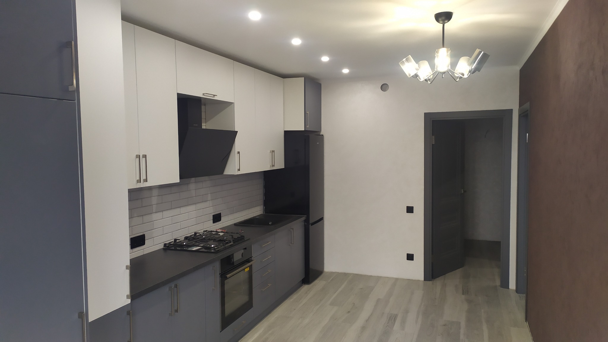 Nowoczesna kuchnia z białymi szafkami górnymi i szarymi dolnymi, czarnym okapem i lodówką, jasnymi płytkami imitującymi cegłę nad blatem, szarą podłogą imitującą drewno oraz dekoracyjną lampą...