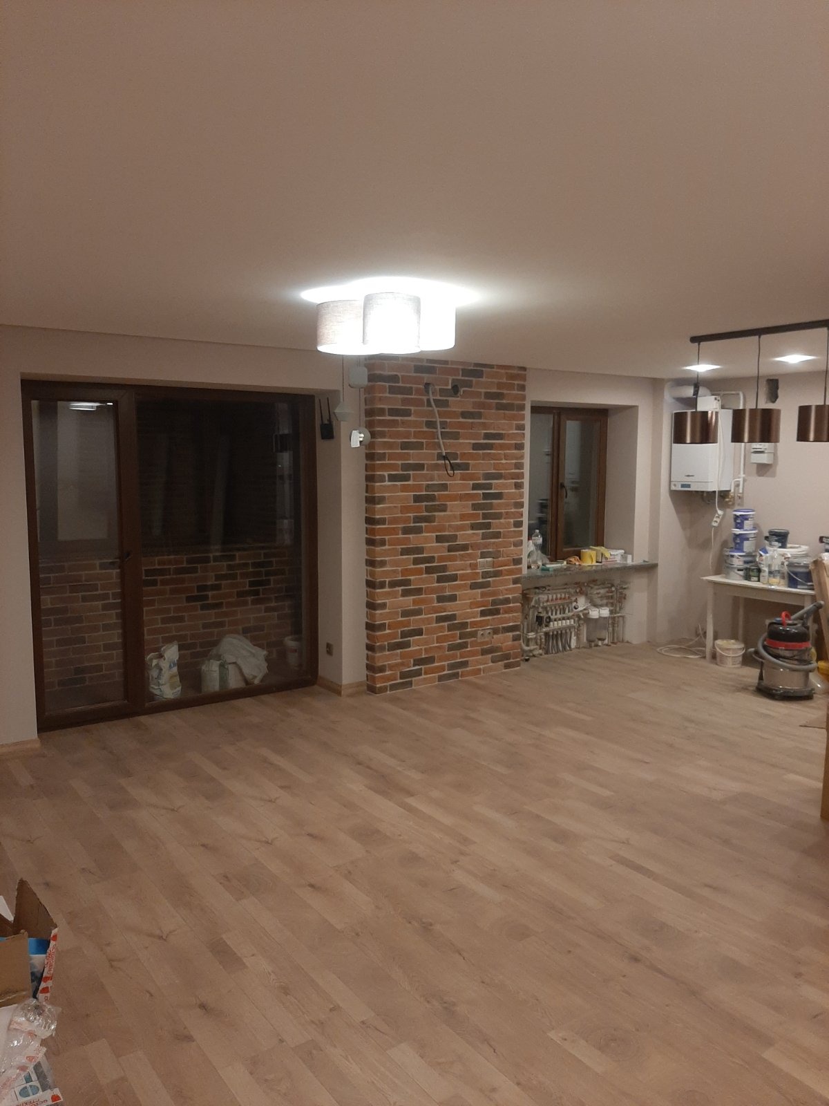 Wnętrze pokoju po remoncie z drewnianą podłogą, ceglaną ścianą i widocznymi instalacjami grzewczymi pod oknem, w tle widoczny sprzęt malarski i odkurzacz przemysłowy.