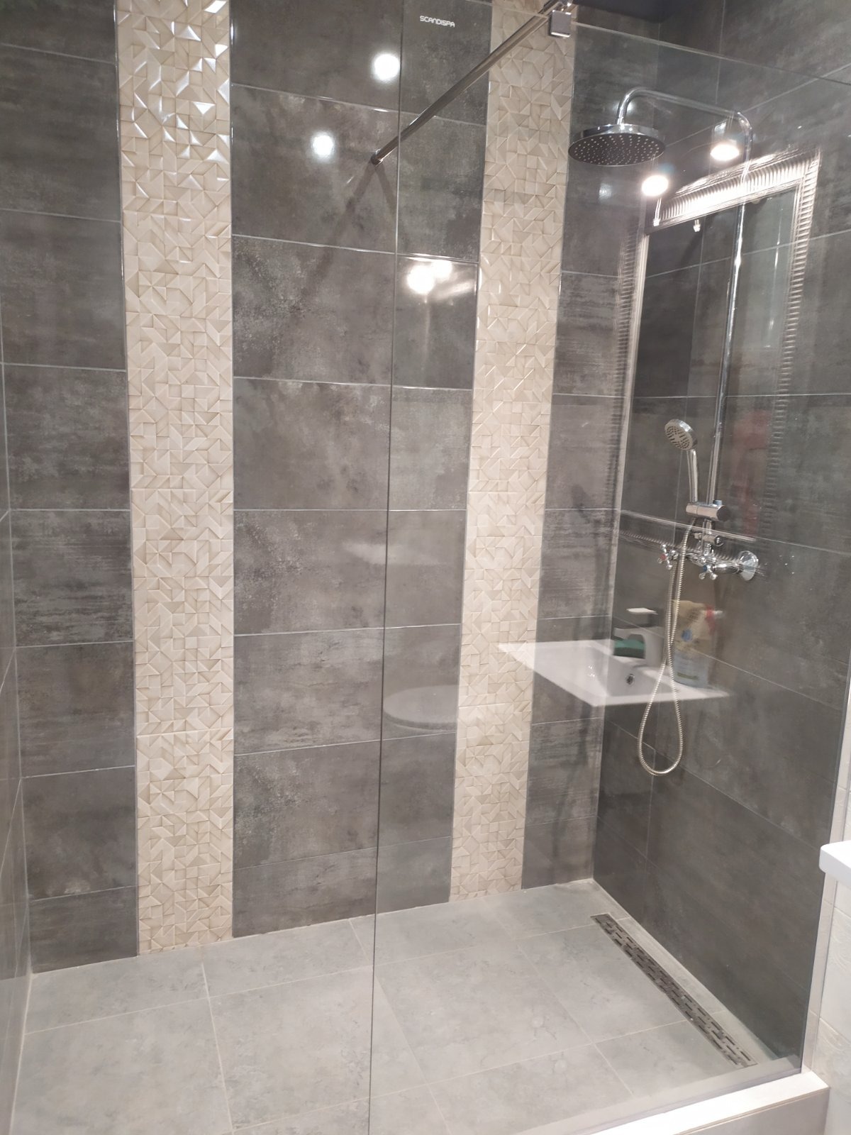 Nowoczesna kabina prysznicowa z szarymi płytkami imitującymi beton i pionowymi pasami z mozaiki w odcieniach beżu, szklana szyba kabiny, deszczownica i bateria prysznicowa.