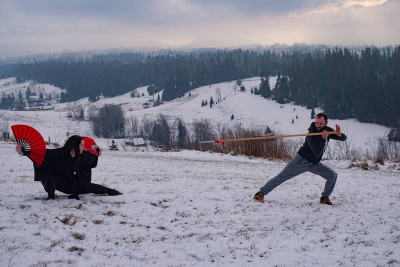 Trening sportów walki na śniegu z użyciem wachlarzy i włóczni na tle górskiego krajobrazu.