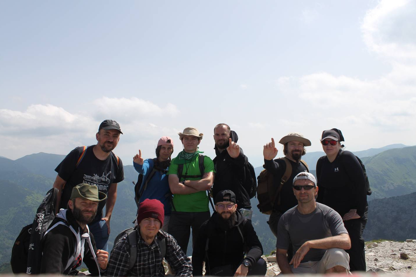 Grupa dziesięciu osób, w różnym wieku i ubraniach turystycznych, pozuje na szczycie góry z widokiem na pasmo górskie w tle, część z nich wskazuje palcem do góry.