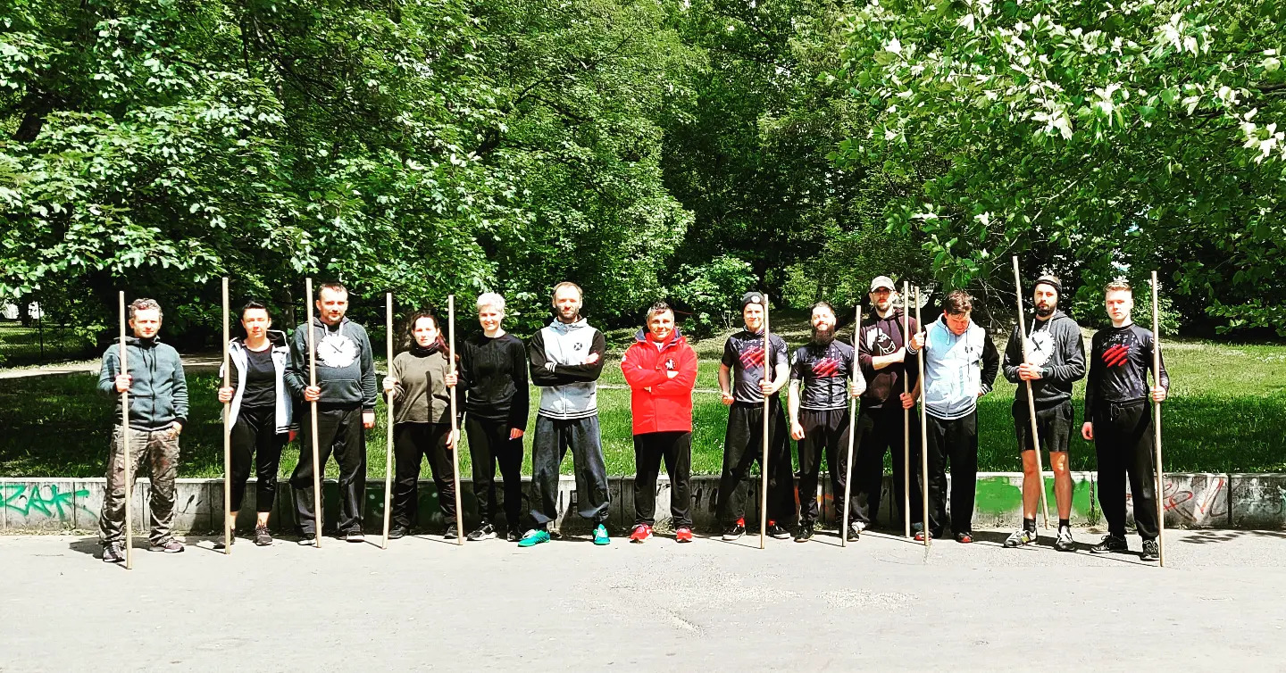 Grupa osób, w tym kobiety i mężczyźni, w strojach sportowych, stojąca na zewnątrz z długimi, drewnianymi kijami, prawdopodobnie podczas treningu sztuk walki z użyciem broni białej na tle zieleni...