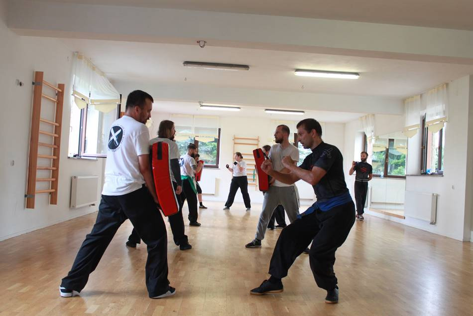 Grupa osób trenuje sporty walki w jasnej sali z drewnianą podłogą; mężczyzna w czarnym stroju z niebieskim pasem demonstruje ruchy, a inni trzymają czerwone tarcze treningowe.