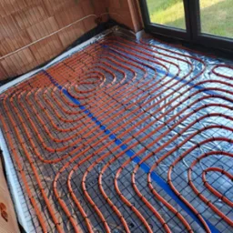 Pomarańczowe rury ogrzewania podłogowego ułożone spiralnie na srebrnej folii izolacyjnej z niebieskimi paskami, widoczne ściany z czerwonej cegły i okno z widokiem na zieleń.