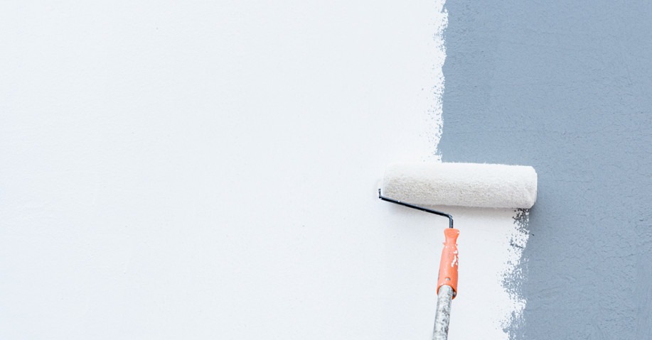 Ściana malowana wałkiem na szaro, kontrastująca z bielą. Widoczna faktura farby i świeże pociągnięcia. Pomarańczowa rączka wałka dodaje akcentu kolorystycznego.