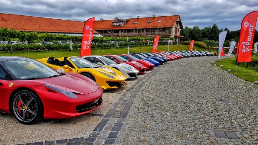 Rząd luksusowych samochodów Ferrari w różnych kolorach, ustawionych na brukowanej drodze przed budynkiem z czerwonym dachem, z flagami Shell V-Power po bokach.