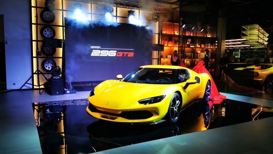 Żółty samochód Ferrari 296 GTB na scenie z reflektorami i wyświetlaczem, częściowo okryty czerwoną tkaniną, widoczne opony na stojaku i osoby w tle.