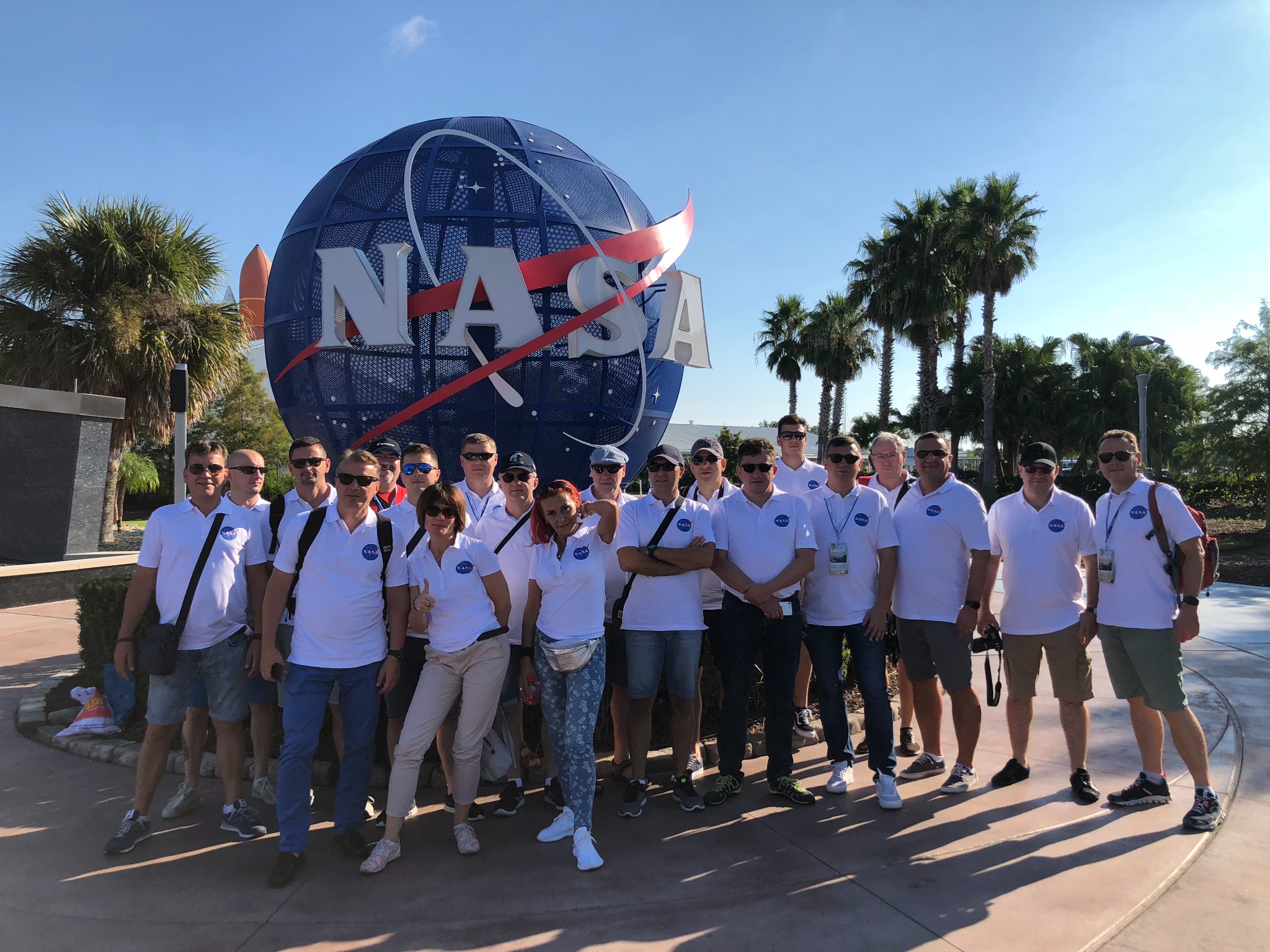 Grupa osób w białych koszulkach z logo NASA pozuje przed dużym, kulistym znakiem NASA na tle palm i błękitnego nieba.