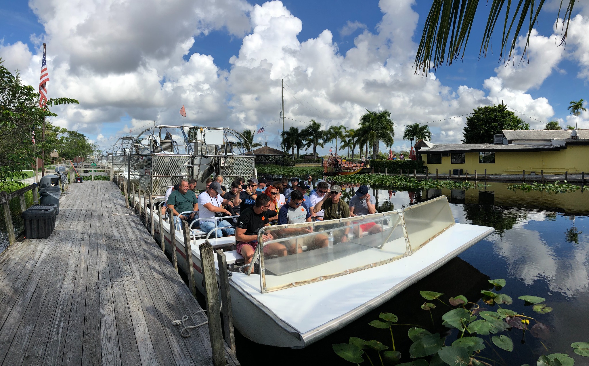 Grupa turystów na pokładzie łodzi motorowej z odkrytym dachem, płynącej po wodzie w otoczeniu roślinności wodnej, z widocznym drewnianym pomostem i budynkami w tle, pod błękitnym niebem z chmurami.