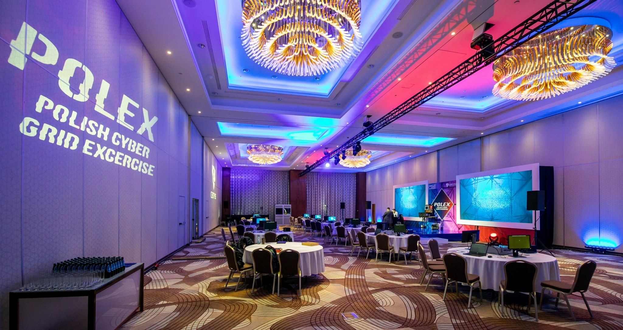 Sala konferencyjna w Warszawie przygotowana na wydarzenie, z okrągłymi stołami, krzesłami, oświetleniem punktowym i projekcją napisu 'POLEX POLISH CYBER GRID EXCERCISE' na ścianie.