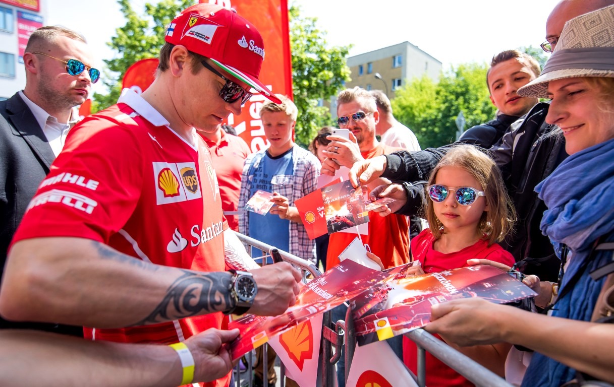 Kierowca wyścigowy w czerwonej koszulce Ferrari z tatuażem na ramieniu, podpisuje kartki dla fanów, w tym dla dziewczynki w okularach z lustrzanymi szkłami.