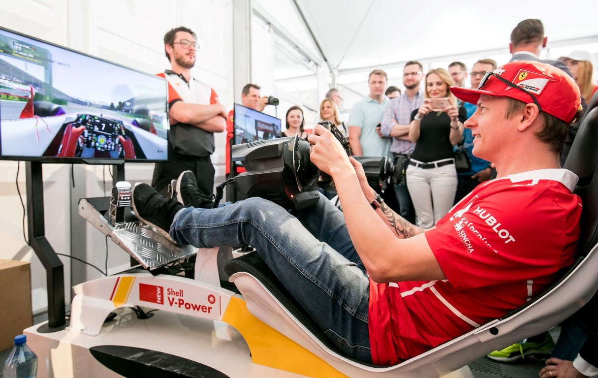 Mężczyzna w koszulce Ferrari i czerwonej czapce z daszkiem, siedzący w symulatorze wyścigów Formuły 1 z wolantem, pedałami i ekranem, otoczony przez obserwujących go ludzi.