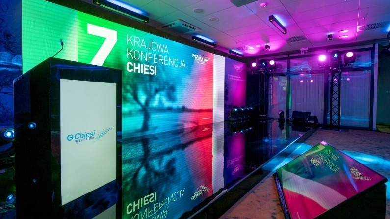 Scena konferencyjna z podświetlanym pulpitem prelegenta z logo firmy Chiesi, duży ekran LED wyświetlający grafikę i napis '7 Krajowa Konferencja Chiesi', oświetlenie sceniczne w kolorach różu...