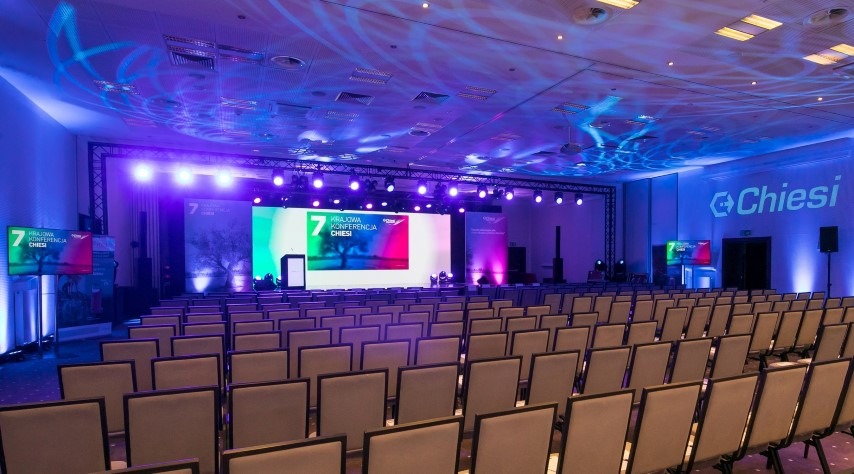 Sala konferencyjna z rzędami krzeseł ustawionych przed podświetloną sceną z ekranami wyświetlającymi logo i nazwę '7 Krajowa Konferencja Chiesi', oświetlenie punktowe na suficie.