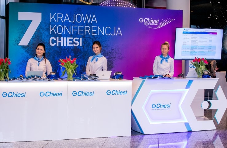 Trzy kobiety w błękitnych apaszkach stoją za białymi stanowiskami recepcyjnymi z logiem Chiesi podczas 7. Krajowej Konferencji Chiesi, z tulipanami w wazonach i monitorami wyświetlającymi...