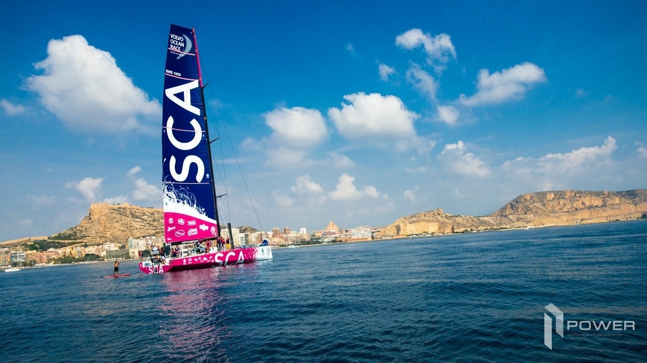 Żaglówka z dużym napisem SCA na żaglu płynie po morzu na tle miasta i wzgórz w słoneczny dzień.