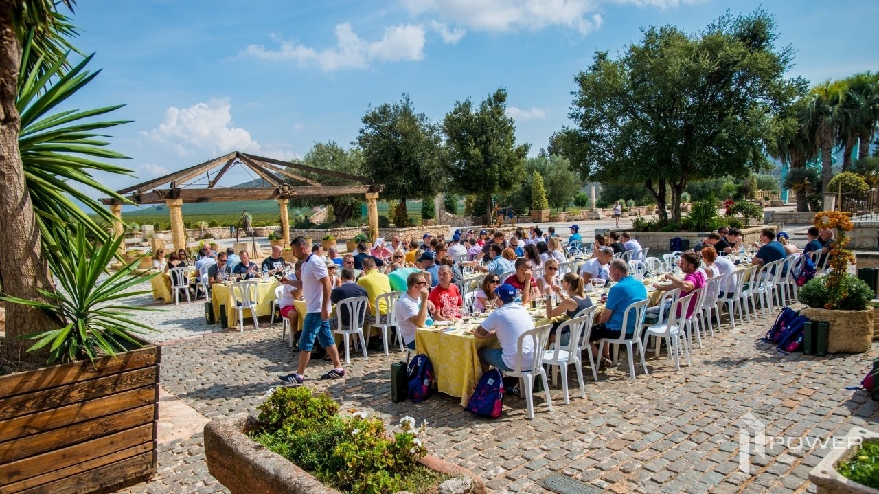 Uczestnicy siedzą przy długich stołach z żółtymi obrusami na zewnątrz, w tle zadaszona konstrukcja i drzewa, na pierwszym planie palma i kostka brukowa.