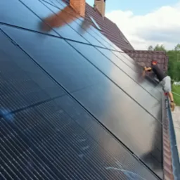 Montaż paneli fotowoltaicznych na dachu domu jednorodzinnego z ceglanym kominem, instalator w okularach ochronnych na drabinie, pochylony nad panelami.