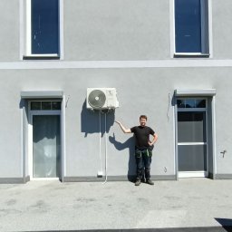 Dobra Energia Jan Filipiuk - Mężczyzna w ubraniu roboczym prezentuje zamontowaną jednostkę zewnętrzną klimatyzacji na szarej ścianie budynku z dwoma oknami na piętrze i dwoma drzwiami na parterze.