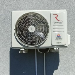 Dobra Energia Jan Filipiuk - Zewnętrzna jednostka klimatyzacji Rotenso zamontowana na szarej, teksturowanej ścianie budynku, z widocznymi białymi rurami odprowadzającymi skropliny i kablami, oświetlona naturalnym światłem...