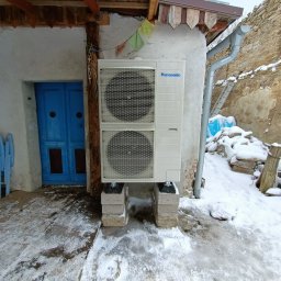 Dobra Energia Jan Filipiuk - Biała jednostka zewnętrzna Panasonic na betonowych bloczkach, zamontowana na ścianie budynku obok niebieskich drzwi, zimowy krajobraz w tle.