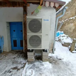 Biała jednostka zewnętrzna Panasonic na betonowych bloczkach, zamontowana na ścianie budynku obok niebieskich drzwi, zimowy krajobraz w tle.