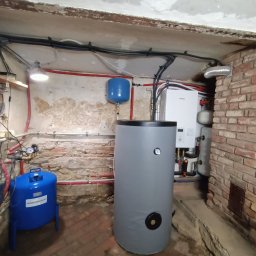 Dobra Energia Jan Filipiuk - Instalacja pompy ciepła w piwnicy: szary zasobnik ciepłej wody użytkowej, niebieski zbiornik hydroforowy, biała jednostka wewnętrzna pompy ciepła Panasonic na tle ceglanej ściany i zniszczonego...