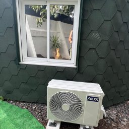 Dobra Energia Jan Filipiuk - Zewnętrzna jednostka klimatyzacji AUX zamontowana na betonowych bloczkach przed domem z zieloną elewacją imitującą gont, obok sztuczna trawa i kamienie.