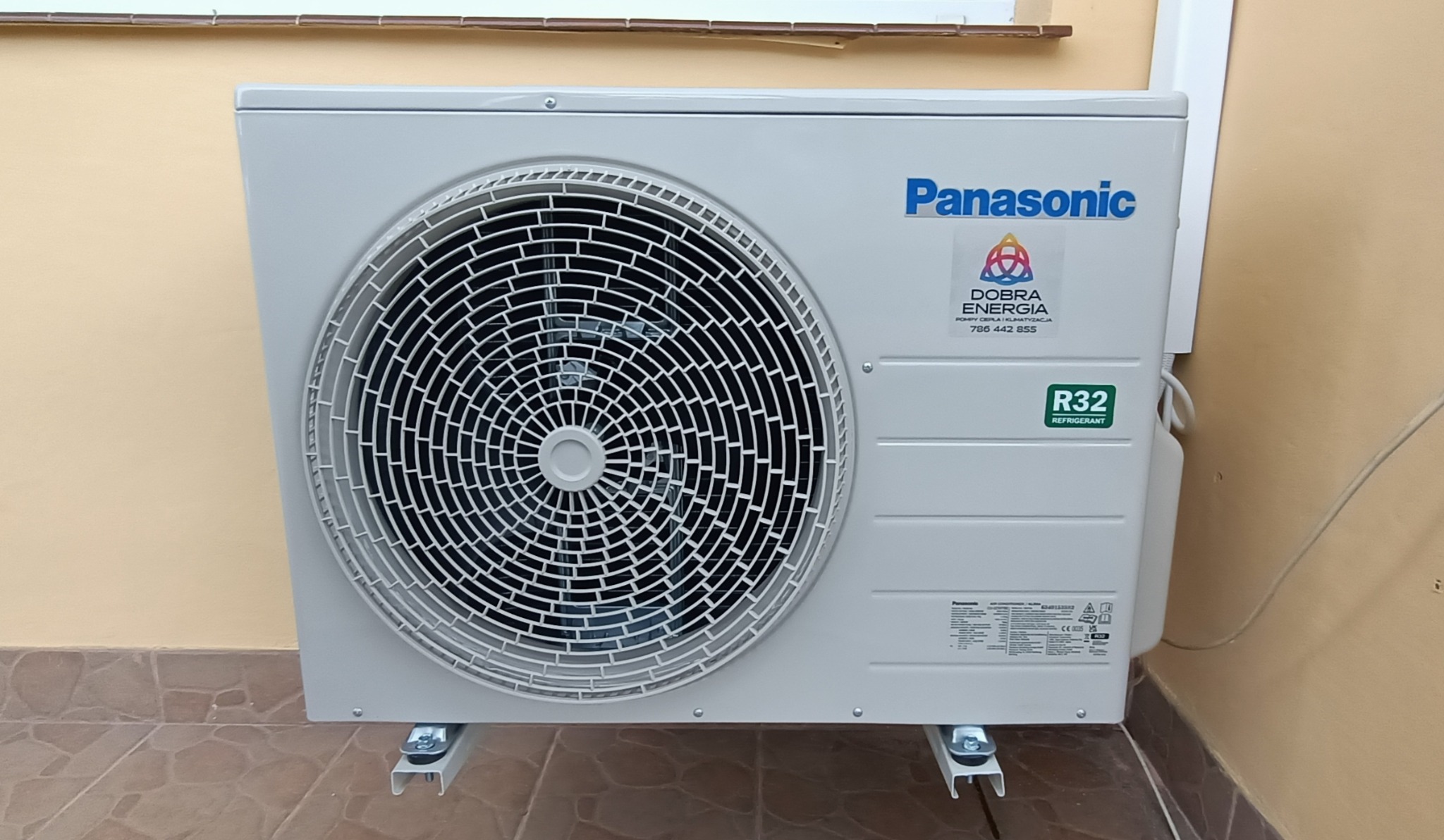 Zewnętrzna jednostka klimatyzacji Panasonic zamontowana na metalowych wspornikach przy ścianie budynku, z naklejką firmy instalacyjnej 'Dobra Energia' i informacją o czynniku chłodniczym R32.