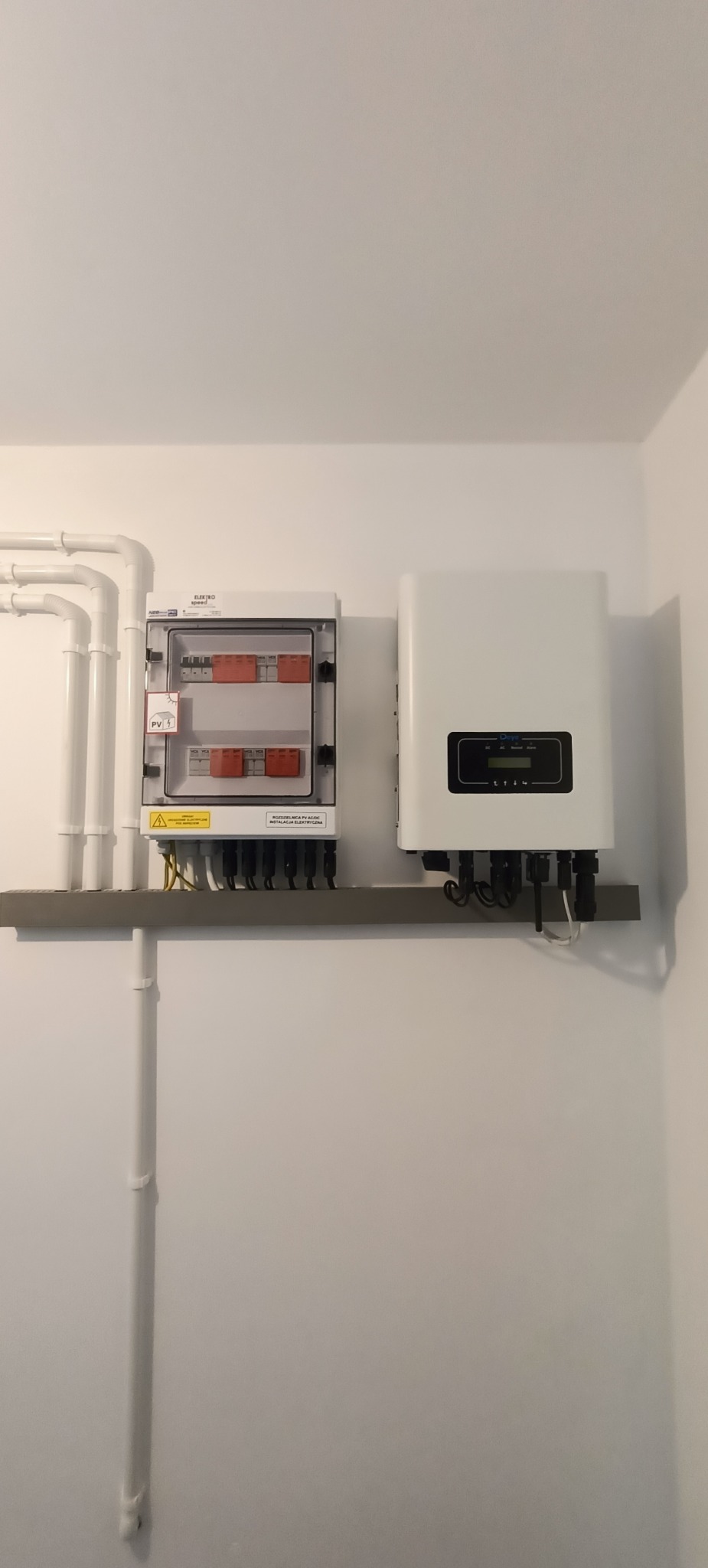 Biała ściana z zamontowaną skrzynką bezpiecznikową z widocznymi bezpiecznikami oraz falownikiem fotowoltaicznym na szarej półce, z widocznymi przewodami elektrycznymi.